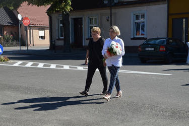 Zdjęcie przedstawia delegację jednostek pomocniczych gminy niosącą bukeit kwiat&oacute;w. Dwie kobiety idą obok siebie. Jedna ubrana na czarno, druga w jasną koszulę i ciemne spodnie.