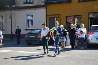 Zdjęcie przedstawia w&oacute;jta Gminy Mieścisko wraz z przewodniczącą Rady Gminy niosoących wiązankę. Mężczyzna ubrany w granatowy garnitur, kobieta białą bluzkę i jasny żakiet oraz zielone spodnie.