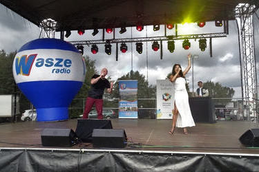 Na zdjęciu widać duet kobietę i mężczyznę. Oboje śpiewają. Z lewej strony znajduje się mężczyzna w ciemnym stroju.Obok niego jest kbieta w jasnej sukience.