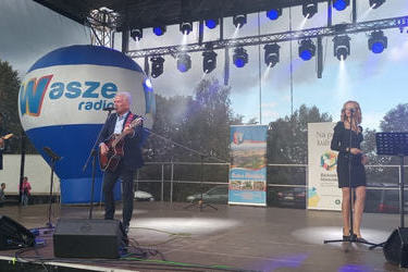 Zdjęcie przedstawia siwego mężczyznę z gitarą i mikrofonem. Obok niego stoi drugi mężczyzna w okularach, r&oacute;znież śpiewa. Z drugiej strony znajduje się kobieta w ciemnej sukience, ma przed sobą mikrofon.
