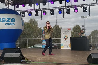 Zdjęcie przedstawia wokalistę na scenie. Ubrany jest w ciemne spodnie i złotą marynarkę.