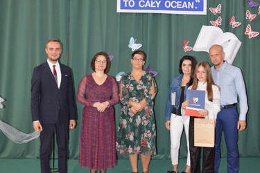 Zdjęcie przedstawia od lewej strony w&oacute;jta, panią dyrektor w okularach i śliwkowej sukni, następnie radna w kwiecistej sukience, obok stypendystka w galowym stroju, za nią stoją rodzice.