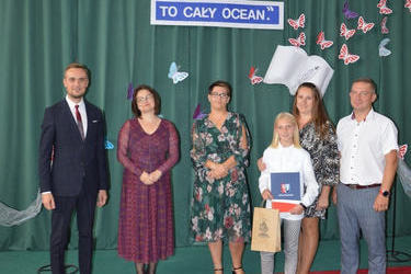 Zdjęcie przedstawia od lewej strony w&oacute;jta, panią dyrektor w okularach i śliwkowej sukni, następnie radna w kwiecistej sukience, obok stypendystka w białej koszuli i spodniach, za nią rodzice.