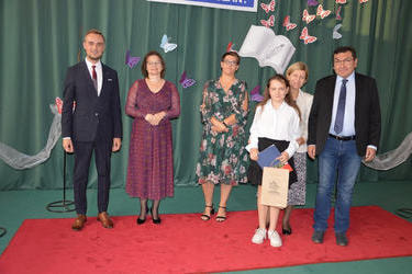 Zdjęcie przedstawia od lewej strony w&oacute;jta, panią dyrektor w okularach i śliwkowej sukni, następnie radna w kwiecistej sukience, obok stypendystka w stroju galowym, z nią stoją rodzice w eleganckich strojach.