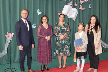 Zdjęcie przedstawia od lewej strony w&oacute;jta, panią dyrektor w okularach i śliwkowej sukni, następnie radna w kwiecistej sukience, obok stypendystka w żakieciku i sp&oacute;dniczce,a za nią mama w czarnych włosach, ciemnym stroju i beżowym żakiecie.