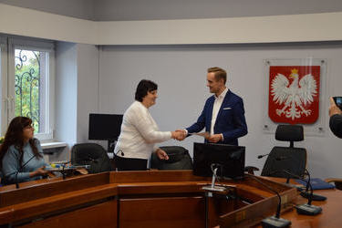 Zdjęcie przedstawia w&oacute;jta gminy podającego dłoń opiekuńce ukraińskiej młodzieży. Kobieta ma kr&oacute;tkie, ciemne włosy, jasny sweter. W&oacute;jt ma granatową marynarkę. Oboje są uśmiechnięci.