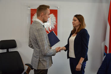 Zdjęcie przedstawia w&oacute;jta wręczającego nagrodę nauczycielce. Kobieta ma brązowe włosy za ramiona, ubrana jest w biała bluzkę i granatowy żakiet  i granatowe spodnie.