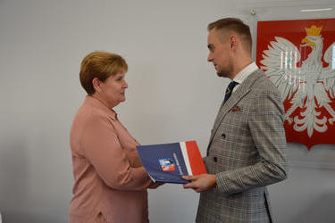 Na zdjęciu w&oacute;jt wręcza nagrodę nauczycielce. Kobieta ma kr&oacute;tkie, brązowe włosy, koszulę w odcieniu r&oacute;żu. W&oacute;jt ma na sobie białą koszulę i marynarkę w kratkę.
