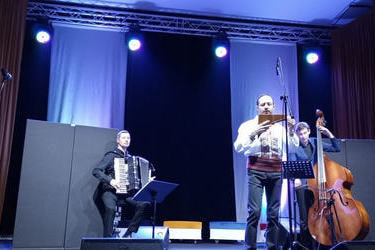 Na zdjęciu widać mężczyznę grającego na instrumencie fletnia pana.Scena oświetlona jest niebieskim światłem. Za grającym mężczyzna znajdują sie inni muzykanci grający na instrumentach.