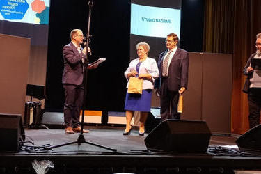 Na zdjęciu znajduje się prowadzący i starsza para. Kobieta w niebieskiej sukni i jasnym żakiecie, obok mężczyzna w ciemnym garniturze.