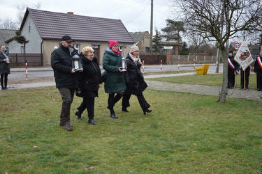 Zdjęcie przedstawia czworo uczestnik&oacute;w podchodzących do pomnika. Dw&oacute;ch z nich niesie znicze.
