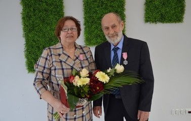 Zdjecie przedstawia parę jubilat&oacute;w. Kobieta w brązowych włosach do uszu. Ma sukeinkę w kratę. Obok mężczyzna w ciemnym garniturze. Ma siwą brodę.