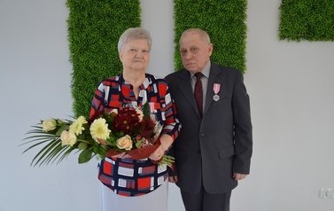 Zdjęcie przedstawia parę jubilat&oacute;w. Kobieta w kr&oacute;tkich, jasnych włosach, ma kolorową bluzkę, w ręce trzyma bukiet kwiat&oacute;w. Mężczyzna ma ciemny garnitur.