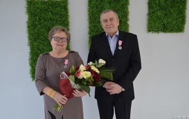 Zdjęcie przedstawia parę jubilat&oacute;w. Kobieta ma kr&oacute;tkie, ciemne włosy, okulary, brązową sukienke i w ręce trzyma bukiet kwiat&oacute;w. Mężczyzna ma ciemny garnitur.