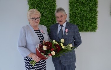 Zdjęcie przedstawia parę jubilat&oacute;w. Kobieta kr&oacute;tkie, jasne włosy i okulary. Ma na sobie błękitny żakiet i trzyma w ręce kwiaty. Mężczyzna w szarym garniturze.