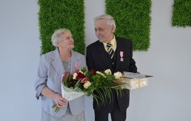 Zdjęcie przedstawia parę jubilat&oacute;w, kt&oacute;rzy patrzą na siebie. Kobieta w siwych, kr&oacute;tkich włosach. Ma na sobie błękitną garsonkę. Mężczyzna w ciemnym garniturze. 