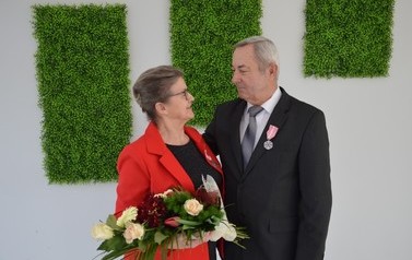 Zdjecie przedstawia parę jubilat&oacute;w, kt&oacute;rzy patrza sobie w oczy. Kobieta w spiętych włosach, czerwonym żakiecie i z kwiatami w ręce. Mężczyzna w ciemnym garniturze.