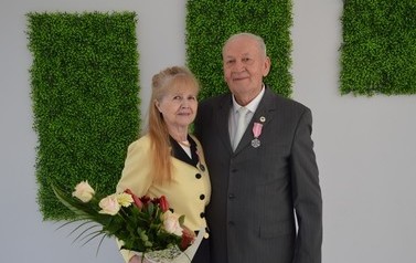 Zdjęcie przedstawia parę jubilat&oacute;w. Kobieta w długich włosach i ż&oacute;łtym żakiecie, trzyma bukiet kwiat&oacute;w. Obok mężczyzna w ciemnym garniturze.