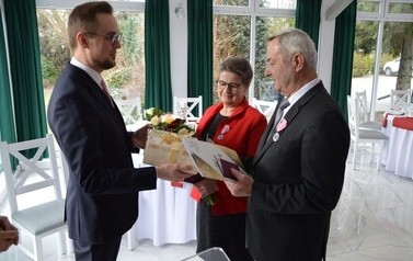Na zdjęciu w&oacute;jt wręcza prezenty jubilatom. Mężczyzna w ciemnym garniturze, kobieta w czerwonej marynarce. 