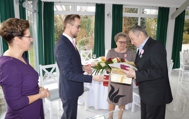 Na zdjeciu w&oacute;jt wręczna podarunki starszemu małżeństwu. Na twarzach wszystkich widac uśmiechy. Wszyscy ubrani są elegancko.