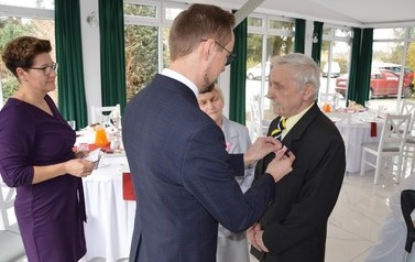 Na zdjęciu w&oacute;jt przypina medal starszemu mężczyźnie. Jest on skupiony. Obok stoi małżonka. 