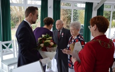 Zdjęcie przedstawia parę jubilat&oacute;w, kt&oacute;rym w&oacute;jt wraz z przewodniczącą wręczają kwiaty i podarunki.Starsze małżeństwo jest skupione. Oboje mają przyczepione medale.