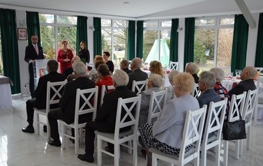 Zdjęcie przedstawia od tyłu jubilat&oacute;w siedzących na krzesłach, przed nimi przy m&oacute;wnicy stoi prowadząca. 