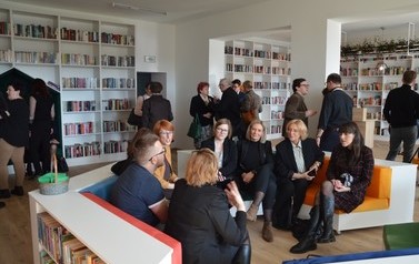Na zdjęciu ludzie podziwiają nowowczesne wnętrze biblioteki. Białe p&oacute;łki z książkami, kolorowe miejsca do siedzenia. 