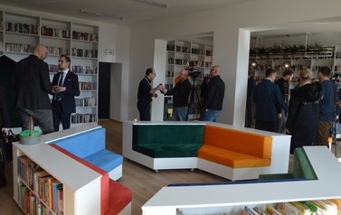 Na zdjęciu ludzie podziwiają nowowczesne wnętrze biblioteki. Białe p&oacute;łki z książkami, kolorowe miejsca do siedzenia. 