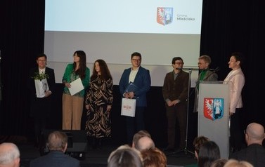 Zdjęcie przedstawia kilka os&oacute;b stojących na scenie, jedna z nich wygłasza przemowę.