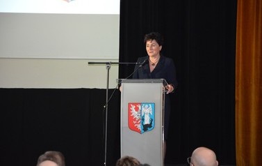 Zdjęcie rpzedstawia przemawiającą kobiete w ciemnych włosach. Stoi na scenie przy m&oacute;wnicy.