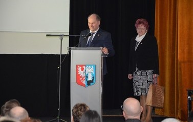 Zdjęcie przedstawia przemawiającego mężczyznę, obok stoi starsza kobieta. W ręce trzyma podarunek.