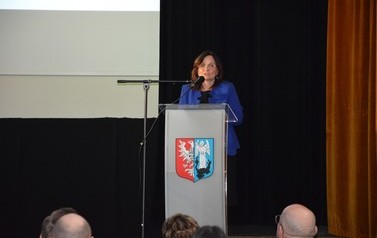 Zdjęcie przedstaiwa kobietę wygłaszajacą przemowę na scenie przy m&oacute;wnicy.