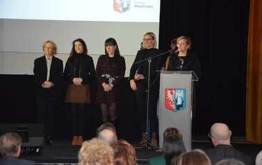 Zdjęcie przedstawia pięć kobiet stojących na scenie. Jedna z nich wygłasza przemowę przy m&oacute;wnicy. 