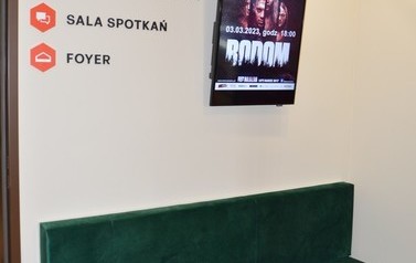 Zdjecie przedstawia korytarz biblioteki. Zielona sofa, a nad nią ekran.
