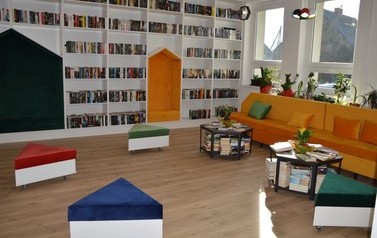 Zdjęcie przedstawia nowowczesne wnętrze biblioteki. Białe p&oacute;łki z książkami, kolorowe miejsca do siedzenia. 