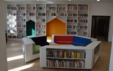 Zdjęcie przedstawia nowowczesne wnętrze biblioteki. Białe p&oacute;łki z książkami, kolorowe miejsca do siedzenia. 