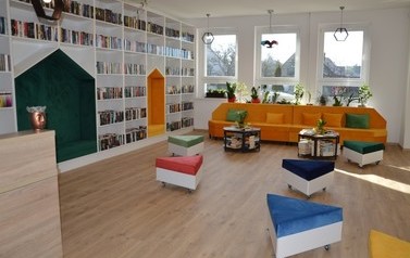 Zdjęcie przedstawia nowowczesne wnętrze biblioteki. Białe p&oacute;łki z książkami, kolorowe miejsca do siedzenia. 