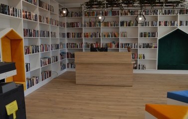 Zdjęcie przedstawia nowowczesne wnętrze biblioteki. Białe p&oacute;łki z książkami, kolorowe miejsca do siedzenia. 