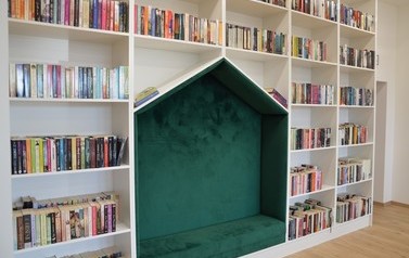 Zdjęcie przedstawia nowowczesne wnętrze biblioteki. Białe p&oacute;łki z książkami, kolorowe miejsca do siedzenia. 