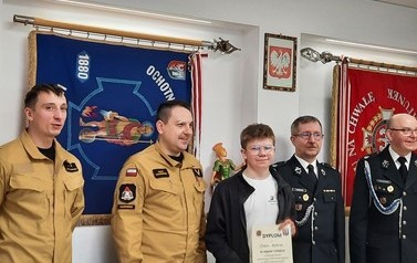 Mężczyźni w strojach strażackich z chłopcem trzymającym w ręku dyplom za zajęcie I miejsca. W tle sztandary strażackie