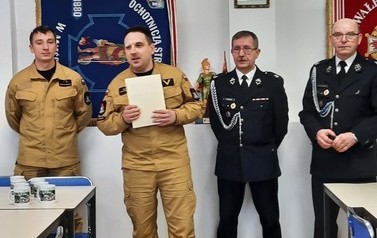 Czterech mężczyzn ubranych w stroje strażackie.Od lewej pierwszych dw&oacute;ch w mundurach straży państwowej. Dwaj kolejni w mundurach wyjściowych . W tle widać sztandary strażackie