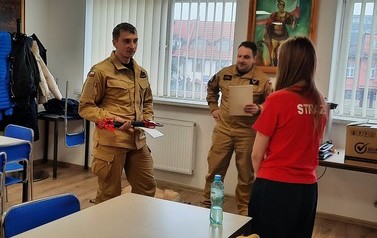 Dziewczyna sto0jaca tyłem przed dwoma mężczyznami ubranymi w stroje strażackie