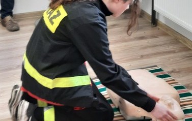 Dziewczyna ubrana w str&oacute;j strażacki pokazująca na fantomie pierwszą pomooc