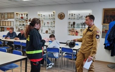 Dziewczynka ubrana w str&oacute;j strażacki opowiada o przedmiocie trzymanym przez mężczynę - strażaka. W tle widać ścianę z pucharami