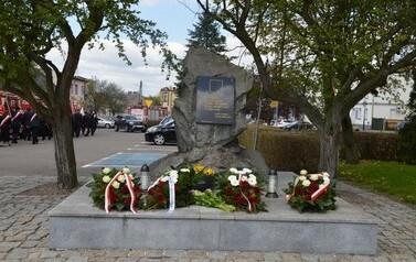 Zdjęcie przedstawia pomnik na kt&oacute;rym znajdują się wiązanki, kwiaty oraz znicze.