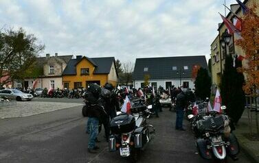 Tyły motocykl&oacute;w oraz grupa mężczyzn ubrana na czarno w kaskach znajduje się przy swoich maszynach, do kt&oacute;rych zotały przyczepione polskie flagi.