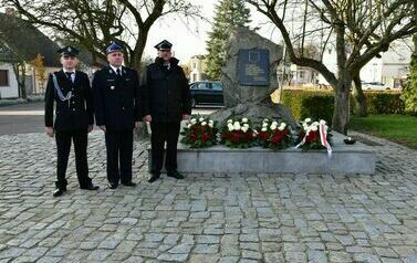 Trzech mężczyzn ubranych w czarne mundury stoi przy pomniku, na kt&oacute;rym znajdują się biało-czerwone kwiaty. 