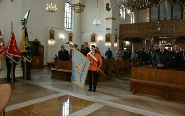 Na środku kościoła stoi chłopak ubrany w pomarańczową kurtkę, trzymający przed sobą sztanadar. W tle widoczni są inni uczestnicy mszy.