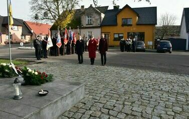 Kobieta ubrana na czerwono oraz dw&oacute;ch mężczyzn ubranych na czarno stoi przed pomnikiem ze spuszczonymi w d&oacute;ł głowami. Za nimi po lewej stronie zdjęcia stoi grupa mężczyzn trzymająca sztandary.